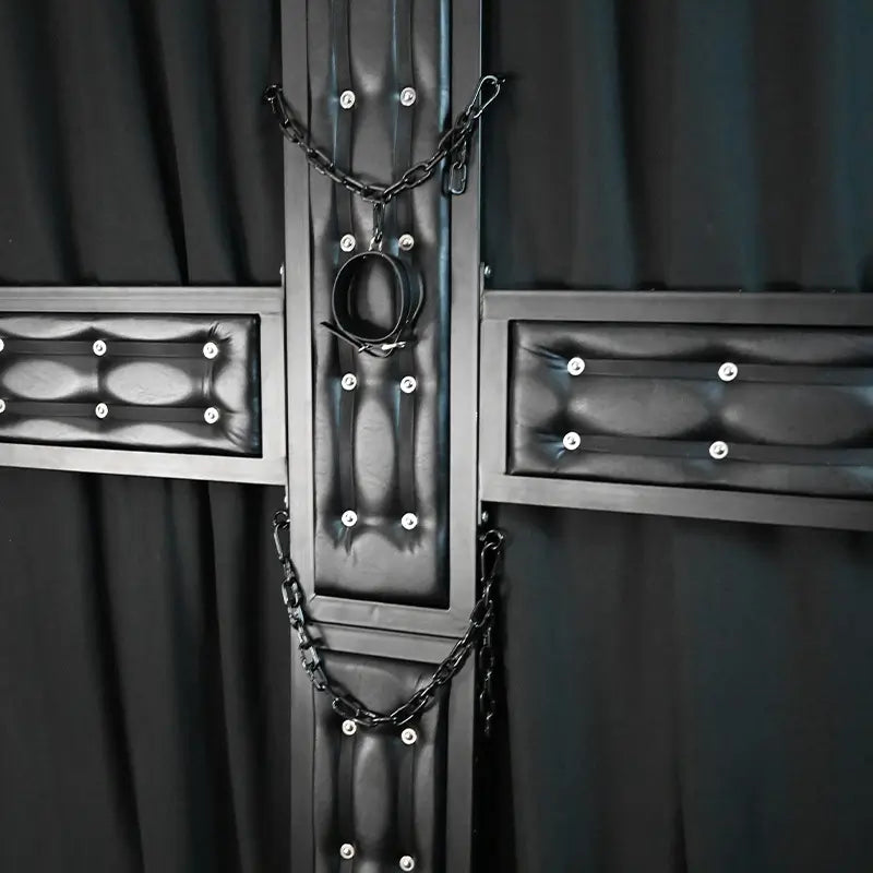 Close-up view of a premium black St. Andrew’s cross featuring padded leather-style panels, reinforced steel frame, restraint chains, and central restraint ring. Luxury BDSM furniture detail designed for upscale dungeon interiors, private playrooms, and commercial studio environments.