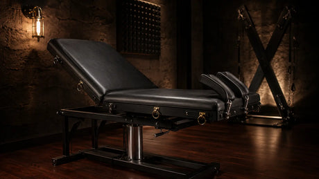 Premium black leather BDSM examination table with brass restraint points and integrated stirrups in a moody dim private studio, BDSM Authority