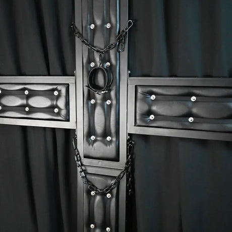 Close-up view of a premium black St. Andrew’s cross featuring padded leather-style panels, reinforced steel frame, restraint chains, and central restraint ring. Luxury BDSM furniture detail designed for upscale dungeon interiors, private playrooms, and commercial studio environments.
